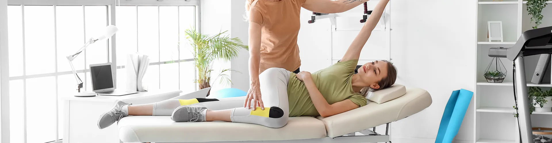 Chiropractor stretching woman’s leg and arm while she lies on her side on therapy bed.