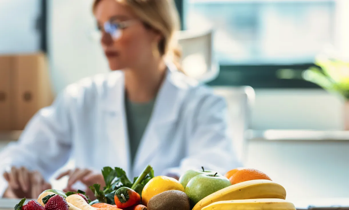 Fruits and vegetables in foreground with blurred practitioner using computer in background.